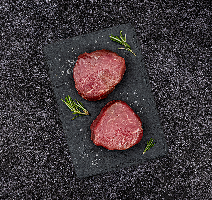Two raw beef steaks on a dark slate board with rosemary sprigs, on a dark textured surface.